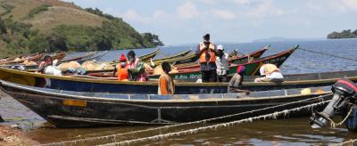 Several wooden boats are moored by a lakeshore, with people wearing life jackets engaged in various activities on and near the boats. The background features hilly terrain and a clear sky.