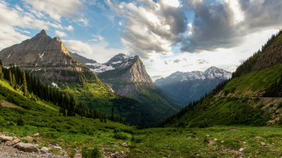 Mountain valley view with grassy slopes, coniferous trees, and snow-capped peaks under a partly cloudy sky. A narrow road is visible on the right hillside.