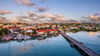 Coastal cityscape with colorful buildings along the waterfront, boats docked at a pier, and a calm sea reflecting the blue sky and scattered clouds.