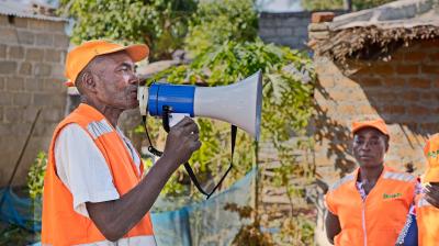 A man in an orange vest and cap speaks into a megaphone outdoors while another person in similar attire stands nearby.
