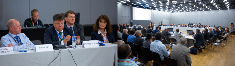 Wide-angle view of a conference room with a panel of speakers and a large audience. Participants sit at a rectangular arrangement of tables with microphones and nameplates.