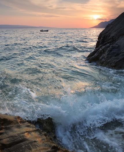 Ocean waves crash against rocky cliffs at sunset, with a small boat visible on the distant water and mountains along the horizon.